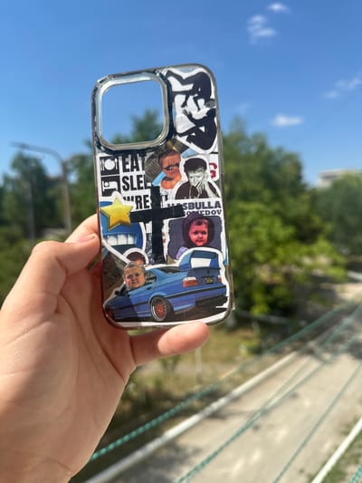 A hand holds a clear phone case adorned with stickers, against a backdrop of a bright blue sky and blurred green trees, suggesting an outdoor setting in Criuleni, Moldova. The stickers feature various images, including a car, a yellow star, and multiple portraits of individuals, one of whom is a child. The phone case is transparent, allowing the hand and the background to be partially visible. The overall lighting indicates a sunny day.