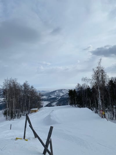 The image displays a snow-covered ski slope within a mountainous landscape in Novoabzakovo, Russia, under an overcast, cloudy sky. The foreground presents a wide, groomed ski path. To the immediate left, several wooden posts forming a broken barrier are partially buried in the snow, with a yellow object visible near their base. Along the left perimeter of the slope, a dense line of deciduous trees with bare branches and dark coniferous trees are visible. Further down this left edge, a small, light-colored building with a dark roof is situated among the trees. In the mid-ground, a prominent snow ramp or jump is sculpted into the slope. A single individual, appearing as a small red figure, is located on the slope past this ramp. The right side of the slope is also bordered by a mix of bare deciduous and coniferous trees. A yellow and red sign, featuring the number "4", is affixed to a pole or tree on this right side. The background consists of multiple layers of snow-covered mountains and hills extending to the horizon under the consistent grey sky.