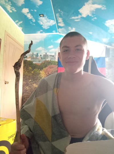 A young male, likely a teenager or pre-teen, with short dark hair and a bare torso, poses for a photo indoors. He looks directly at the camera with a slight smirk. In his left hand, he holds a long, gnarled wooden stick or branch. He is partially covered by a blanket featuring a geometric pattern in shades of grey, yellow, and white.
The room exhibits distinct wallpaper designs: one wall features a mural depicting an autumnal forest landscape transitioning into a cityscape, while an adjacent wall is covered in blue wallpaper adorned with white clouds, resembling a sky. The ceiling appears light yellow or pale green. In the upper left background, a portion of a large yellow bag is visible, bearing Russian text that reads "НДЕКС" (part of "Yandex"). A light-colored door or cupboard is also seen. Bright light illuminates the scene, suggesting it is daytime. An object with red, white, and blue stripes, possibly a flag or fabric, is visible in the lower right background. The setting is identified as совхоза "Воронежский", Russia.