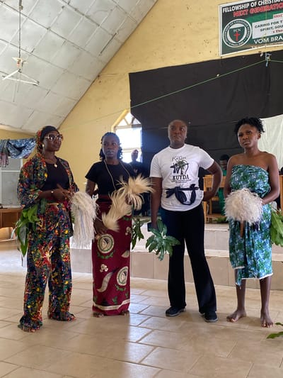 The image shows a group of four women standing indoors, possibly at a church or community hall in Dutsen Kato, Nigeria. The women are of African descent and appear to be participating in a cultural or religious event.
The woman at the front is wearing a colorful, patterned outfit with a headscarf and sunglasses. She holds leafy branches in her hands. Next to her, a woman in a black shirt and a maroon, patterned skirt is also holding leafy branches and a white, fluffy object. The third woman, in a white t-shirt with text and black pants, stands to the left of the maroon-skirted woman. The woman at the back, wearing a blue and green patterned strapless dress, holds a similar white, fluffy object. The overall mood seems festive and ceremonial.
Visible text on a banner in the background reads: "FELLOWSHIP OF CHRISTIAN NURSES NEXT GENERATION", "FCN", "Kufere", "KUYDA", "FUL DEVELOPMENT ASSOC.", "1 Thess. 5:23", and "BEROM WOMEN DEVELOP..."
The indoor setting has tiled floors and walls, and a black backdrop is visible behind the women. Natural light streams in from above, suggesting it is daytime. The presence of leafy branches and traditional attire hints at a local cultural celebration or ceremony.