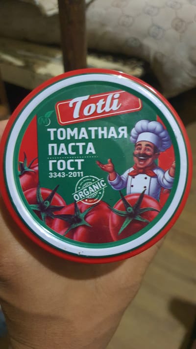 The image shows a close-up of a person's hand holding a round can of tomato paste. The can has a red, white, and green striped border. The label on the can features the brand name "Totli" in red lettering within a white box. Below this, in white Cyrillic letters, is "ТОМАТНАЯ ПАСТА," which translates to "Tomato Paste." Beneath that, also in white, is "ГОСТ 3343-2011." There are graphics of tomatoes and a chef character wearing a chef's hat and a red bowtie, with his arms outstretched. To the left of the chef, there is a circular "ORGANIC" seal. The background of the can is green, and the images of tomatoes are red. The hand holding the can is brown-skinned. The environment beyond the can is blurry, with hints of a wooden surface and fabric. The city of Karankul, Uzbekistan, is not depicted in the image.