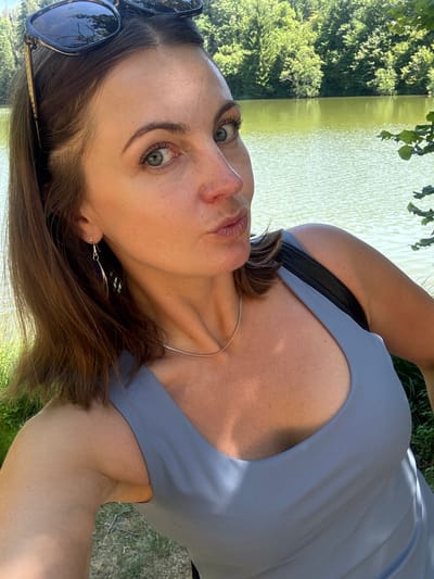 A woman with brown hair and green eyes, wearing sunglasses, a grey tank top, and long dangle earrings, is taking a selfie outdoors near a body of water. She is puckering her lips and looking at the camera. In the background, there are trees and a lake. The image is a close-up shot, with the woman's face and upper body filling most of the frame. The woman appears to be on a dirt path next to the lake, with grass and foliage visible on the left and right sides of the image. The scene takes place in a natural environment, likely a park or forest. It is not possible to determine the specific location within Ravilloles, France from the image.