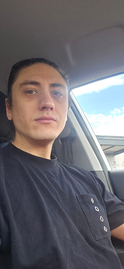 A close-up, medium shot captures a person from the chest up, seated in the passenger seat of a car. The individual, with dark hair pulled back, is looking directly at the camera. They are wearing a dark blue t-shirt with a pocket adorned with three silver grommets. The background shows a bright blue sky with scattered white clouds, suggesting an outdoor setting. Part of the car's interior, including the window frame and seatbelt, is visible. The location is identified as Bous, Germany.