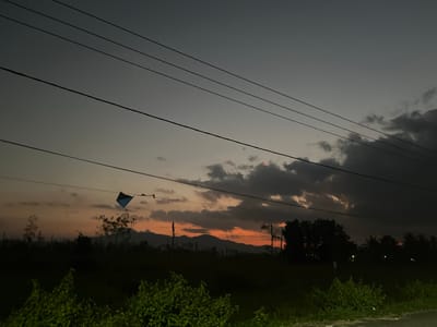 The image depicts a serene rural landscape at dusk, with power lines stretching across the frame. In the middle ground, a kite, predominantly blue with some black accents, is suspended from a wire. Beyond the kite, a dark silhouette of trees and vegetation lines the horizon, with a faint glow of the setting sun visible behind a range of mountains in the distance. The sky transitions from a pale grey at the top to shades of orange and dusky purple near the horizon, indicative of twilight.

The scene suggests a peaceful evening in a rural area, possibly a field or open space. The presence of power lines implies proximity to a settlement or infrastructure, while the natural elements like the trees, mountains, and the kite suggest a location conducive to outdoor activities. The overall mood is tranquil and contemplative, perhaps evoking a sense of calm as day transitions to night. The time of day is clearly evening, as indicated by the dramatic sky colors and dimming light. There is no visible text in the image. The location context is Batujai, Indonesia.