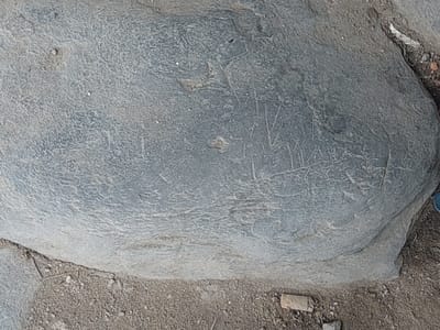 The image shows a large, gray, weathered rock with some faint markings etched into its surface. The rock is positioned on the ground, which is covered with dirt, small stones, and some debris. The environment appears to be outdoors, possibly in a natural or semi-developed area within the city of Parit Baru, Indonesia, given the context provided. There are no people visible in the image. The markings on the rock are indistinct, appearing as thin, scratched lines, possibly representing symbols or inscriptions, though their exact nature is not clear. The lighting suggests it is daytime, and the overall scene is one of an aged, static object in a simple outdoor setting.