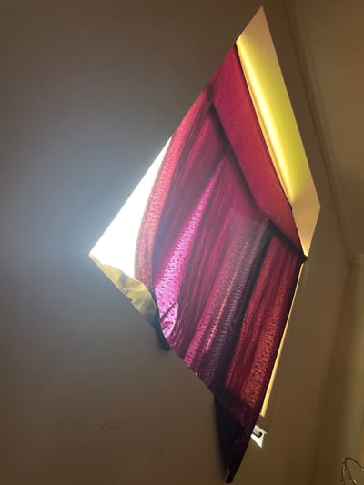 The image shows a close-up, angled view of a triangular-shaped window or skylight, with a portion of a dark red curtain visible. The window is illuminated from the outside by bright sunlight, causing the curtain to be translucent in some areas and casting shadows. The interior walls are painted a light beige color. The angle of the shot suggests the viewer is looking up towards the ceiling. The scene is devoid of any people or discernible urban environment. Given the request to include a location in Hazorasp, Uzbekistan, and the fact that the image itself does not contain any identifying features of that city, it is impossible to provide this detail based solely on the visual information. The image depicts an interior architectural detail with natural lighting.