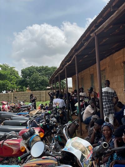 The image depicts an outdoor scene in Foram, Nigeria, likely a public gathering. In the foreground, a row of parked motorcycles, some red and one white, dominates the frame. One motorcycle displays a poster with the image of a man and text that reads "GONE TOO SOON!" and "Late Mr. GWOM JERRY JOHN".

Behind the motorcycles, a covered, open-air structure with a corrugated metal roof is visible, supported by wooden posts. The structure appears to be made of brick. Several people are gathered under this shelter, some sitting and others standing. They are dressed in a variety of clothing, including patterned fabrics and a plaid shirt. One woman in the center, wearing a black headscarf and dress, is holding what appears to be a phone and taking a picture. Another woman is wearing a colorful headscarf and patterned dress.

In the background, a brick wall is visible, and beyond that, lush green trees suggest a vegetated environment. The sky above is partly cloudy with white and grey clouds. The overall impression is a community gathering, possibly a memorial or event, with motorcycles serving as transportation and a central point of focus.