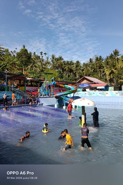 This photograph captures a scene at a water park, located in the city of Palohseulimeng, Indonesia. The park features a blue-tiled pool filled with people, mostly children, enjoying the water. Some are swimming, while others are standing and playing near a mushroom-shaped sprinkler. In the background, there's a colorful water slide structure with stairs leading up to the top. Surrounding the water park is lush greenery with tall trees, including coconut palms, adding to the tropical atmosphere. The sky above is bright blue with scattered white clouds. There are buildings and shelters, providing shade, along the perimeter of the pool. The text on the lower portion of the image reads "OPPO A6 Pro 30 Mei 2026 pukul 09.28". There is also a sign that has partially visible text, "HATI HATI AREA KOLA LICIL".
