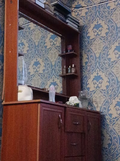 The image depicts an interior scene, likely a room in an apartment or house, possibly located in Кызыл-Октябрь, Kyrgyzstan. The dominant feature is a dark wood vanity or dresser with drawers and shelves. On the top shelf, there are several books, a white object that appears to be a baby bottle or a similar container, and a few small bottles. To the right of the vanity, the wall is covered with patterned wallpaper in blue and cream colors. The lighting appears to be dim, contributing to a slightly grainy quality of the image.