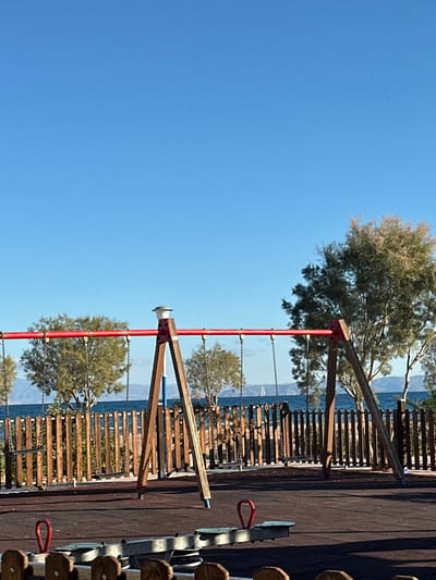 This image depicts a children's playground situated by the sea in Megalochori, Greece, under a clear, bright blue sky. The foreground shows a dark reddish-brown rubberized safety surface. A brown wooden seesaw with red handles is partially visible in the bottom-center. Dominating the mid-ground is a red-beamed swing set featuring two brown wooden A-frame supports with blue-tipped bases; a white cap-like fixture tops the left support. Two swing chains hang from the red crossbar. A wooden picket fence, consisting of vertical slats, separates the playground from the sea. Beyond this fence, the deep blue water of the sea is visible, with scattered green-leafed trees along the coastline. In the distant background, hazy blue-grey landmasses rise above the horizon. A small white sailboat with a single sail is discernible on the water to the right. No individuals are present in the scene.