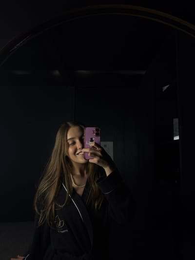 A young woman is taking a mirror selfie in a dark room. She is wearing a black robe with white trim and a pearl necklace. She is holding a pink smartphone with stickers on it in front of her face. The mirror has a dark frame and reflects the dimly lit room, partially revealing architectural details. The location is suspected to be in Ciudad López Mateos, Mexico, but there are no visible clues within the image to confirm the precise location.