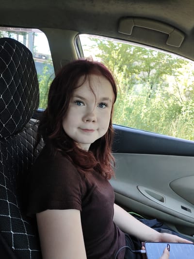 A young woman with reddish-brown hair and blue eyes sits in the passenger seat of a car. She is wearing a dark brown ribbed shirt and has her right arm resting on her lap, holding a smartphone. The car's interior is visible, including the black seat cover with a white diamond pattern on the left and the door panel with a gray armrest on the right. Outside the car window, lush green foliage and trees are visible under a bright sky. The scene is set on a sunny day, suggesting an outdoor location, possibly a road trip or a stop during travel, likely in or near Shimanovsk, Russia, given the car's interior and the surrounding vegetation characteristic of the region.