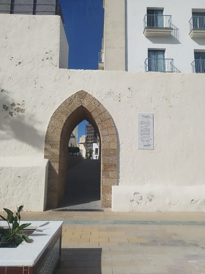 This is a medium shot, taken outdoors on a sunny day, of an ancient stone archway leading into a narrow, paved pathway. The archway is made of rough-hewn, tan-colored stone blocks. The wall surrounding the arch is whitewashed and shows signs of age and wear, with some plaster peeling away.

To the right of the arch, mounted on the whitewashed wall, is a plaque with handwritten text. The text appears to be a poem or a historical inscription written in a dark ink.

Beyond the archway, the pathway slopes gently upwards and disappears into the distance. A few buildings are visible in the background, including a modern white building with balconies and windows. Scaffolding is visible on one of the distant buildings, suggesting ongoing construction or renovation.

In the foreground, to the left of the arch, is a raised planter box with a few green plants. The ground in front of the arch and planter is paved with interlocking bricks in various shades of yellow, beige, and gray, with a thin green stripe running horizontally across the paving.

The clear blue sky and the strong shadows cast by the wall and the archway indicate that it is daytime, likely mid-morning or afternoon. The overall scene evokes a sense of history and timelessness, with the ancient archway juxtaposed against modern architecture in the background. There are no people visible in the image. The location is Rota, Spain.