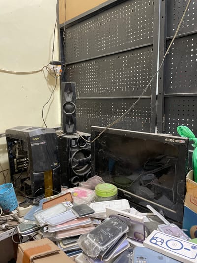 The image depicts a cluttered desk or workstation in what appears to be a workshop or storage area in Pasar Paloh, Indonesia. The scene is dominated by electronic equipment and various accessories. In the background, a large pegboard structure is visible, with wires hanging down.
Prominently featured is a black computer tower case, covered in dust and cobwebs, to the left of a two-piece speaker system. The speaker system consists of a taller, thin speaker unit stacked on top of a larger speaker unit. A black monitor, also dusty, is positioned to the right of the speakers, with a green plastic bag partially obscuring its right side. The monitor screen shows signs of damage or cracking.
In the foreground, a significant pile of mobile phone cases is arranged on the surface. These cases vary in color and material, with some still in their original packaging. Other small electronic accessories and miscellaneous items are scattered amongst the phone cases. A blue plastic organizer is visible to the far left. A cardboard box is partially visible at the bottom right corner. The overall impression is one of disarray and neglect, possibly indicating a repair shop, an electronics recycling area, or a storage space for unsold inventory.