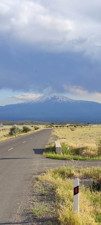 A winding asphalt road leads through a grassy plain towards a snow-capped mountain under a cloudy sky. The road, marked with white lines, has a shadow cast on its left side. On the right side of the road, a white post with a red reflector stands amidst dry grass. In the distance, a range of hills can be seen, with the imposing mountain dominating the horizon. The sky is filled with dark, heavy clouds, but breaks of light suggest the sun is present. Two birds are visible in the sky, one closer to the mountain and another further away to the left. The overall environment suggests a rural or semi-rural setting, possibly near Lanjazat, Armenia, given the distinctive mountain landscape.