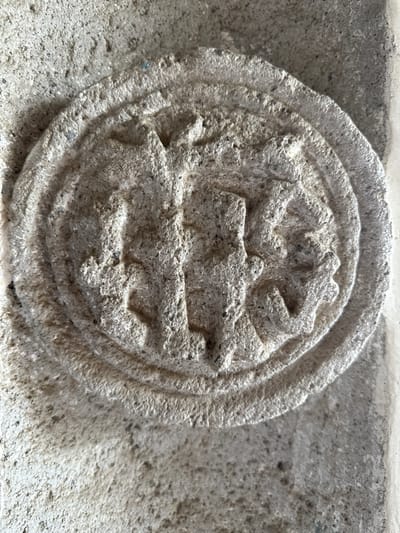 A close-up, overhead shot captures a circular, bas-relief carving made of light-colored stone, embedded in a rough, textured stone wall. The carving appears weathered and displays a central, indistinct figure or symbol with circular elements around it. The wall itself is composed of a coarse, speckled material, with lighter and darker shades of gray. The lighting casts shadows that highlight the texture and depth of the carving and the surrounding wall. This artifact is located on the facade of the Ex-Convento Agustino de San Miguel Arcángel in Huejotzingo, Mexico.