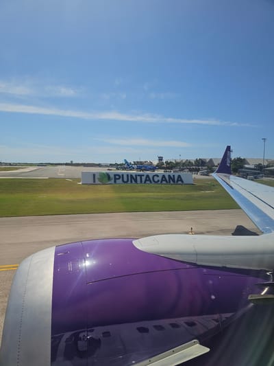 From an airplane window, a partial view of the wing and fuselage of an Arajet airplane in Punta Cana, Dominican Republic. Outside, a sign reads "I <heart> PUNTA CANA". The sky is blue with some clouds.