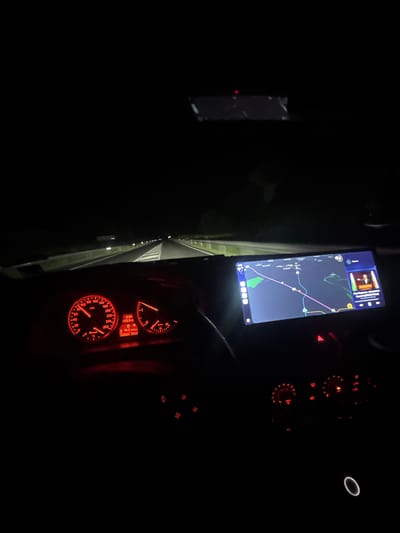 The image shows the interior of a car at night, likely while driving. The speedometer and other dashboard gauges are illuminated in red. A navigation app is displayed on a screen mounted near the dashboard, showing a map with a route marked in purple. The location is likely in Mogila, Bulgaria based on the map displayed on the navigation app. The image captures a dark environment with focus on the driver's view of the road and navigational aids.