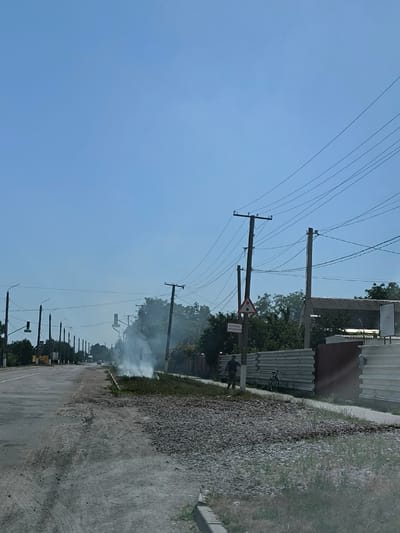 The image displays a daytime street scene in Illintsi, Ukraine, under a clear blue sky. A two-lane asphalt road, showing signs of wear and featuring loose gravel patches on its surface and shoulder, extends from the foreground towards the background on the left. A prominent plume of white smoke rises from ground level on the right side of the road, within a grassy median strip or shoulder area that contains loose gravel.
Multiple wooden utility poles with numerous overhead power lines and cables are visible, traversing the scene from left to right. Streetlights are present along the left side of the roadway. On the right, a dark-clothed individual, appearing male, walks away from the camera, partially obscured by a utility pole. A bicycle is propped against a corrugated metal fence section located behind the walking individual. A triangular road warning sign depicting two vehicles and a rectangular white sign with the Cyrillic text "БЕРЕЖИСЬ АВТОМОБИЛЯ" (Beware of Cars) are affixed to a utility pole on the right. Trees and a building with a dark roof are partially visible beyond the fences on the right side of the frame.
