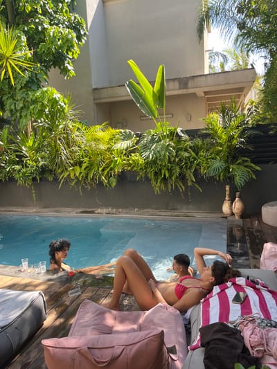 The scene depicts an outdoor leisure area at a private residence, reportedly located in Binyamina - Givat Ada, Israel, under bright daylight conditions. A rectangular swimming pool with clear blue water occupies the midground, surrounded by light-colored wooden decking.
Three individuals are present in or around the pool. In the water, to the left, a female with dark curly hair and darker skin is partially submerged, facing right, holding a clear plastic cup containing a red liquid. To her right, a male subject with short dark hair and lighter skin is also partially submerged, facing left. A second male subject is visible further right in the pool, partially submerged. Multiple clear plastic cups, some containing liquids, are placed on the pool's edge.
In the foreground, to the right of the pool, a female with dark hair and light skin is reclined on her left side on a grey patterned cushion, wearing a red two-piece swimsuit. Her left arm is bent with her hand near her head. A red and white striped towel, a mobile phone, and articles of clothing (a pink top and dark fabric) rest adjacent to her on the cushion. Two beanbag-style cushions, one pink and one grey, are in the lower foreground. An empty clear plastic cup rests on the wooden decking near the pink beanbag.
The background features an elevated, dark-colored planter wall supporting a dense arrangement of various green tropical and subtropical plants, including large-leafed species such as *Monstera* and what appears to be *Strelitzia* (bird of paradise), alongside ferns and other foliage. Behind this verdant display, a light-colored multi-story building with a visible overhang is illuminated by direct sunlight. Two decorative, rustic-style terracotta vases stand on the wooden decking to the right of the pool, next to a small grey rounded pouf.