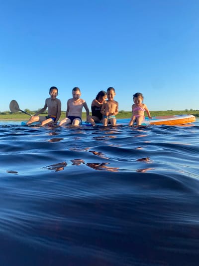 This is a low-angle, vertically oriented photograph taken from the water's surface, looking towards a group of people on what appears to be a paddleboard or raft. The water is a deep blue with ripples and reflections, suggesting a clear day. In the background, a thin strip of green vegetation separates the water from a bright blue sky.
The subjects are a group of six individuals, mostly children and young teenagers, all wearing swimwear. One person at the back is holding a paddle, and another is seated with legs extended. The others are seated or kneeling on the raft. A young girl in a pink bikini is in the foreground, on an orange and white board. The overall atmosphere is one of summer recreation and enjoyment. The lighting suggests it is daytime, likely late afternoon given the long shadows cast by the vegetation.
There is some visible text on the black swimsuit of one of the individuals: "NICE". The location is noted as Bethyuntsy, Russia.