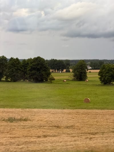 An overcast sky looms over a rural landscape in Kopaniny, Poland. In the foreground, a field of dry, golden stubble stretches horizontally across the image. Behind this, a lush green meadow dominates the middle ground, dotted with three cylindrical hay bales. Further back, a dense line of mature trees forms a dark green border, punctuated by a white building with a dark roof. The background is filled with more trees and the continuation of the sky. The scene appears calm and pastoral, with no people or animals visible.