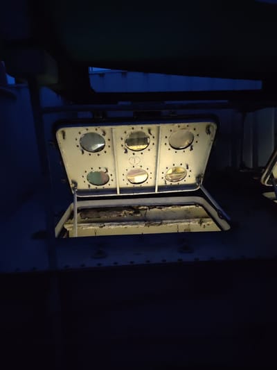 An open hatch with a metal frame, revealing a dark interior space. The hatch has six circular glass windows, some of which reflect light. The frame is attached to a larger metal structure that appears weathered and industrial. The scene is dimly lit, with the light source seemingly coming from within the hatch. The overall environment is dark and appears to be outdoors at night. The location is Badozhskoi Pogost, Russia.