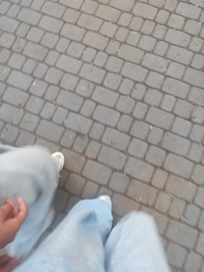 An overhead perspective captures a person's legs and feet walking on a paved surface. The paving stones are rectangular and arranged in a repeating pattern. The person is wearing light-colored pants and white sneakers. A hand is visible on the left side of the frame, appearing to be in motion. The scene is blurred, suggesting movement. The image location is identified as Zhovti Vody, Ukraine.