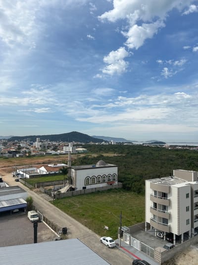 The image is an aerial view of a coastal city in Palhoça, Brazil, under a partly cloudy sky. In the foreground, a modern apartment building with beige and white facades and balconies faces a street. To the left, a light-colored van is parked next to a building with a blue awning. Across the street, a white building with a minaret and arched windows, resembling a mosque, is visible. Further in the distance, a sprawling urban landscape with residential and commercial buildings stretches towards a large, green, tree-covered mountain. The background features a hazy body of water, likely the Atlantic Ocean, with islands and mountains in the far distance. The overall scene depicts a blend of urban development and natural scenery in a Brazilian coastal town.