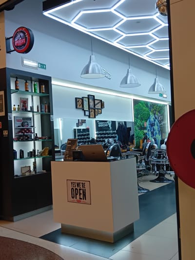 The image shows the interior of a barbershop. There are barber chairs and mirrors visible towards the back. A white, square front desk features a sign reading "YES WE'RE OPEN". The interior is well-lit with a hexagonal lighting fixture on the ceiling. There are shelves displaying products. Various framed pictures are hanging on the wall. The presence of Portuguese signage (the emergency exit sign) and the "Upper Cut" sign visible in the product displays suggest this business is located in Portugal. The specific reference to Amadora is based on the shop name and the presence of the "Upper Cut" shop in Amadora.