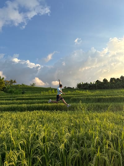 A full shot shows a person in a rice paddy in Padangbulia, Indonesia, on a bright, partly cloudy day. The person, likely male, wears a light-colored t-shirt, dark shorts, and white sneakers. They stand on one leg with arms extended, suggesting a moment of balance or perhaps a pose. The rice paddy stretches into the distance, with neatly organized terraces of green rice plants. A line of trees and foliage is at the top of the photo, marking a transition to denser vegetation. Above, a partly cloudy sky is visible. The image is taken from a low angle, creating a sense of depth. The lighting suggests it is late afternoon, with a warm glow on the rice plants.