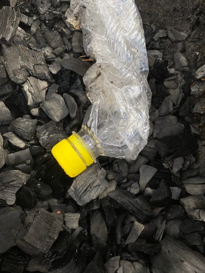 This is a close-up, overhead shot of a clear, crushed plastic bottle with a yellow cap lying on a bed of black charcoal. The bottle is mostly transparent, allowing the texture of the charcoal beneath to be seen. The charcoal pieces are of varying sizes and shapes, some appearing to be rough and jagged, while others have a more splintered texture. The overall scene is dark due to the color of the charcoal, with the bright yellow cap of the bottle providing a stark contrast. There are no people visible in the image. The context provided indicates the location as Leninsky, Russia. No text is visible in the image. The lighting appears to be natural, suggesting it could be daytime. The scene conveys a sense of discarded waste, possibly in an outdoor setting where a fire has recently been extinguished.