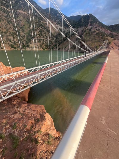 View of the Catwalk Bridge at the Cavallo Dam in Tabaroucht, Morocco. The bridge, with a wood and metal structure, spans over a body of green water, flanked by a red and white painted barrier along the dam. The bridge leads towards a mountainside covered with sparse vegetation under a partly cloudy sky.