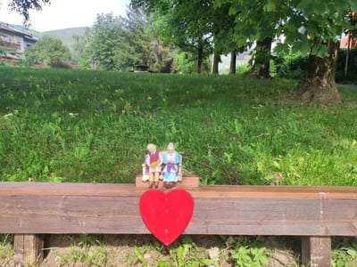 In a grassy outdoor setting, likely a park or garden, a wooden bench is prominently featured in the foreground. Adorning the bench is a bright red heart-shaped decoration. To the right of the heart, two small figurines are seated on a miniature bench. The figurine on the left depicts an elderly man wearing a red vest and white shirt, holding a small cat. The figurine on the right is an elderly woman in a blue dress, knitting with purple yarn. Behind the bench, a lush green lawn stretches out, dotted with scattered branches and leaves. In the background, trees with dense foliage create a natural backdrop. Beyond the trees, buildings and the base of a hillside are visible, suggesting an urban or suburban environment within a mountainous region. The overall scene evokes a sense of peaceful domesticity and affection.