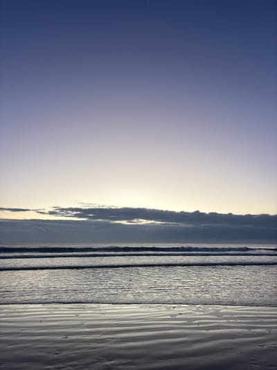 The image captures a serene coastal landscape in Boca del Río, Mexico, at either dawn or dusk. The sky, occupying the upper two-thirds of the frame, exhibits a gradient from a deep indigo hue at the top to a pale yellow-white at the horizon. A faint, white linear contrail is visible in the upper left quadrant of the sky. A horizontal band of dark, layered clouds spans the horizon. Below these clouds, the ocean surface displays multiple parallel wave crests advancing towards the viewer. The immediate foreground consists of wet, reflective beach sand, showing distinct ripple patterns from receding water and subtle indentations, possibly footprints. No individuals, man-made structures, or specific objects are present, and no human activity is depicted.