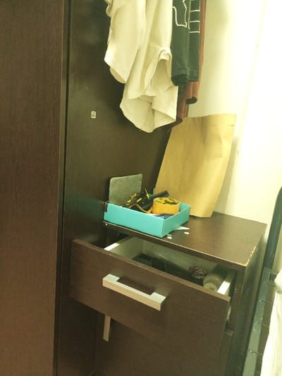 A partially open brown dresser features a silver handle. A light blue box on top contains yellow and black electrical tape, a gray rectangular object, and other items. Hanging above are multiple layers of clothing in white, black, and brown. A large tan paper bag leans against the wall. Items within the drawer include what appears to be a clear plastic bag and a white container.