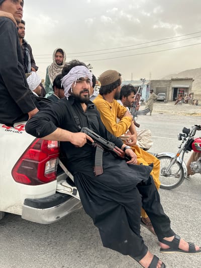 The image depicts a group of armed men lying in the bed of a white pickup truck in Spezand, Pakistan. Most of the men are wearing traditional clothing, including long tunics and head coverings. Several are holding AK-47 style assault rifles. A motorcycle is visible parked nearby. The setting appears to be an outdoor street in an urban or semi-urban area. Buildings can be seen in the background. The weather appears overcast.