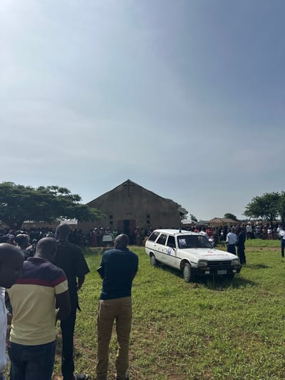 The image captures an outdoor gathering of numerous people in a grassy area under a bright, clear sky, likely during daytime. The scene appears to be set in Panyam, Nigeria.
In the foreground, the backs of several men are visible, indicating a large crowd extending into the background. One man wears a dark blue shirt and khaki trousers, while another has a striped polo shirt and jeans.
A prominent feature in the mid-ground is a white car, clearly marked as an "AMBULANCE" with red lettering across the front and side, accompanied by a red cross emblem. On its side door, the letters "C.H.P" and the word "UTELPINE" are visible. The front license plate displays "AB 565 HKK". The ambulance appears to be an older model sedan, possibly a Peugeot.
Behind the ambulance and crowd, a simple, grey building with a steep, gabled roof stands. It features several rectangular openings towards the top of its facade. The ground is covered in green grass, with some trees visible in the distance and near the building, contributing to the open-air setting. The clear, bright blue sky suggests good weather.
The gathering, centered around the ambulance, suggests a community event, possibly related to public health services, a presentation, or a communal assembly.