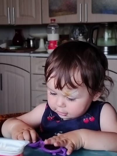 The image shows a close-up of a young child, likely a toddler, sitting at a table in what appears to be a kitchen or dining area. The child has dark hair and is wearing a dark blue sleeveless top with a cherry pattern. A bandage is visible on the child's forehead. The child is looking down and appears to be engaged with something on the table, with their hands reaching out. In the foreground, a portion of a white container and a purple object, possibly a toy, are visible. The background shows kitchen cabinetry, a bottle of liquid, and an electric kettle. The lighting suggests it might be daytime. The general atmosphere is domestic and informal.