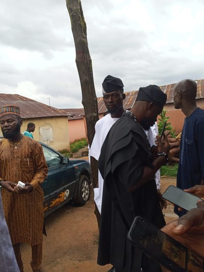 In Panyam, Nigeria, under an overcast sky, a group of men in traditional attire gathers outdoors. The scene is set against a backdrop of residential buildings with corrugated metal roofs. In the foreground, a man wearing a patterned brown tunic and a decorative cap holds a white object in his hands. To his right, another man, dressed in a black robe with layered, structured sleeves and a matching black cap, is looking down at a smartphone he holds. To the left of him, a man in a white robe and black cap is partially visible. Further back, another man in a dark blue tunic is seen. A dark green car with a flame decal is parked behind the man in the brown tunic, and a large tree trunk stands prominently between the man in the black robe and the man in the blue tunic. The ground is covered in reddish-brown dirt.