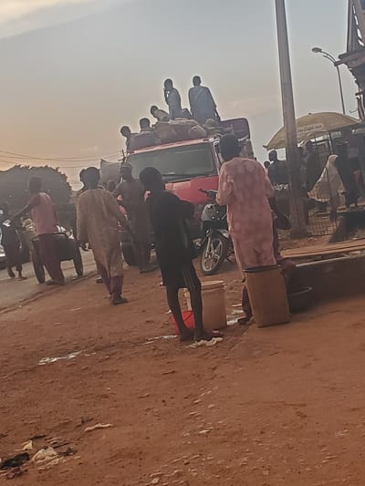 The image depicts a crowded street scene, likely in the city of Funtua, Nigeria, during what appears to be either early morning or late afternoon due to the soft lighting. In the foreground, several individuals are visible. A person in a pink garment is standing next to a large, light-colored barrel, with another barrel and a dark basin nearby. To their left, a young person in a black shirt and shorts stands with their feet in what seems to be shallow water or mud. Further left, a person in a pink shirt and dark pants is pushing a cart with two wheels. Behind them, a person in a tan garment walks away from the camera, and another person in dark clothing is also present.
Dominating the middle ground is a red truck, heavily laden with goods, possibly bags of produce or other merchandise. Several people are perched on top of the truck, some appearing to be communicating or observing their surroundings. One person is speaking on a mobile phone. Another individual, dressed in blue, is seated on the truck.
In the background, a yellow umbrella, advertising "Glo Café app" and "VOICE," is visible, suggesting a commercial area. A street light and the side of a building are also present. The ground is unpaved and appears to be dirt or compacted earth, with some scattered debris and puddles. The overall impression is one of a bustling, active street in a Nigerian urban environment.