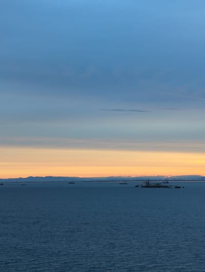 This image depicts a serene seascape at dusk, likely taken from a vantage point in Yokkaichi, Japan, with the Ise Bay stretching towards a distant mountainous horizon. The sky transitions from a deep blue at the top to soft hues of pale orange and yellow near the horizon, indicative of sunset. Several vessels are visible on the water. A cluster of ships and a barge, possibly involved in construction or dredging, are centrally located. Two smaller boats are further to the left, and another ship is visible in the distance on the right. The distant shoreline features a subtle outline of mountains and what appear to be port facilities or city lights. The water itself is a deep blue, reflecting the twilight sky.