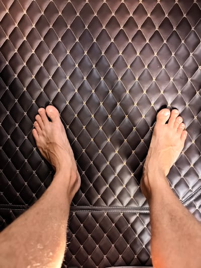 The image shows a pair of bare human feet standing on a dark brown, diamond-patterned surface with light-colored stitching. The surface appears to be a textured mat or upholstery. The lighting suggests an indoor setting. There are no specific landmarks or elements in the image that would indicate a location within Saint-Augustin-des-Bois, France.