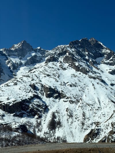 This image shows a snow-covered mountain range against a clear blue sky. The mountain has jagged peaks and steep slopes with a mix of snow and exposed rock. Vegetation is sparse, with patches of trees and shrubs visible near the base of the mountain. A road or path is visible along the right edge of the frame. The location is likely in Villar-d'Arêne, France, given the mountainous terrain typical of the region. No people, objects, or interactions are visible in the immediate scene.