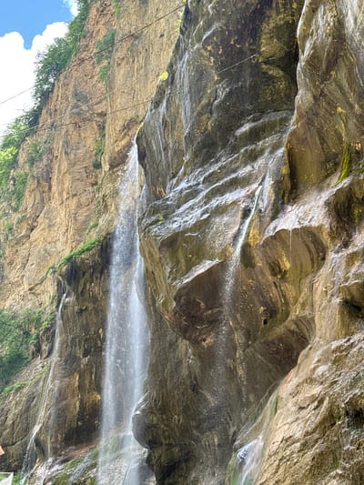 A wide shot captures a dramatic waterfall cascading down a sheer, rugged cliff face. The water, appearing as a thin, luminous stream, descends from an unseen source high above, splashing against the textured rock. The cliff itself is composed of layered stone in shades of brown and gray, with patches of green moss clinging to its damp surfaces. Vegetation, including trees with dense foliage, grows from the top and along the edges of the cliff, contrasting with the starkness of the rock. The sky above is a vibrant blue, with a few wispy white clouds. The overall scene is one of natural beauty and geological grandeur. The location is Khushto-Syrt, Russia.