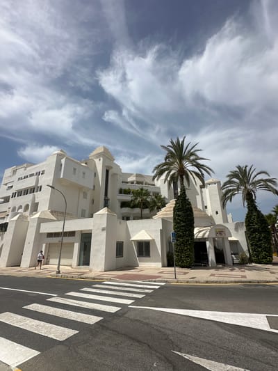 A white, multi-story building with a modern architectural style is visible on a street in Torremolinos, Spain. The building has various geometric shapes, including stepped roofs and a tiered central section. Several palm trees, some covered in ivy, are planted in front of the building. A pedestrian crossing with white stripes is painted on the asphalt street in the foreground. A lone person wearing a white shirt and red shorts is walking on the sidewalk next to the building. The sky above is partly cloudy with wispy white clouds against a blue background. A street lamp stands on the sidewalk near the building. A sign on the building reads "Castillo San Luis."