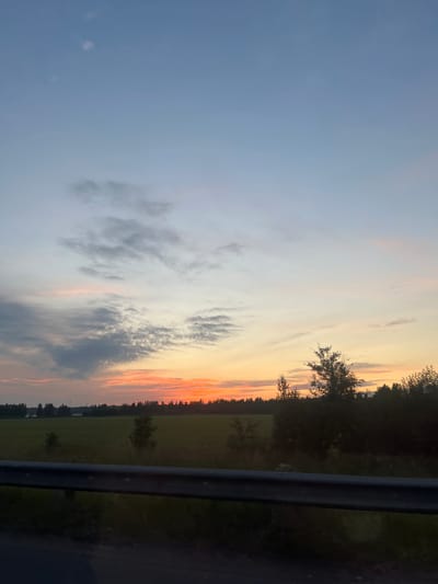 A sunset scene over a rural landscape in Khryastovo, Russia, is depicted. The sky transitions from a pale blue at the top to hues of orange and pink near the horizon, with scattered clouds. Below the sky, a dark line of trees stretches across the middle ground, separating a green field from the sky. In the foreground, a dark, horizontal railing runs across the bottom of the image, obscuring the lower portion of the landscape. The overall impression is one of a peaceful, natural environment at dusk.