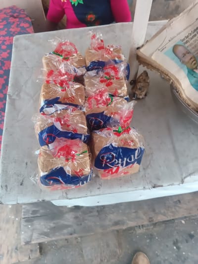 Packaged loaves of bread, labeled "Royal," are arranged on a white, worn table. Each loaf is wrapped in clear plastic and tied with red and green string. Behind the bread, a person wearing a pink top with a navy blue and black floral pattern is partially visible. An old newspaper featuring a person's face is displayed next to a small rock. A single shoe is subtly visible at the bottom of the frame. The overall scene suggests a market setting.