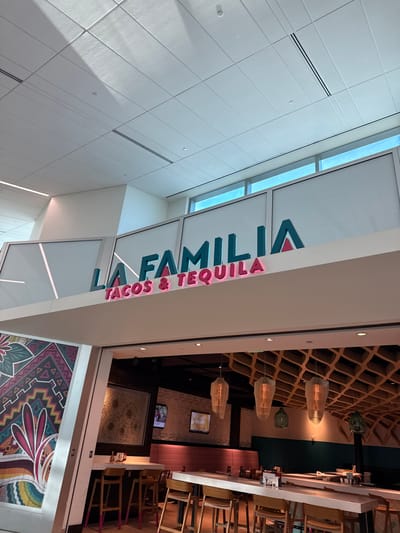 A restaurant called "LA FAMILIA TACOS & TEQUILA" is visible in this image, located in Fort Lauderdale, United States. The restaurant has a modern design with a white ceiling featuring fluorescent lighting and air vents. The signage for the restaurant is prominently displayed in teal and pink. Below the signage, the interior of the restaurant is visible, showing a bar area with wooden stools and tables. The decor includes a colorful mural on the left wall and a unique wooden lattice ceiling with pendant lights in the background. The lighting is a mix of natural light coming from windows and artificial light from the ceiling and pendant lamps. The scene appears to be an interior shot of a commercial establishment.