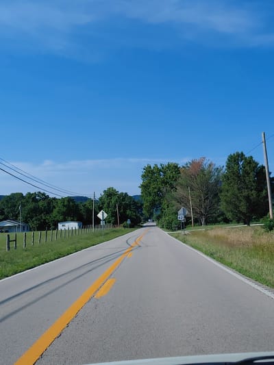 The image depicts a bright, sunny day with a clear blue sky. A paved road, marked with a double yellow line, runs diagonally through the frame, bordered by a grassy verge and trees on the right. On the left side of the road, a sign with "SOUTH 421" is visible, indicating a route number. The scene appears to be rural or suburban, with hints of residential buildings and a white fence line. Power lines also run across the scene. The time of day is likely daytime, given the strong sunlight and blue sky. The image captures a serene and ordinary roadside view.